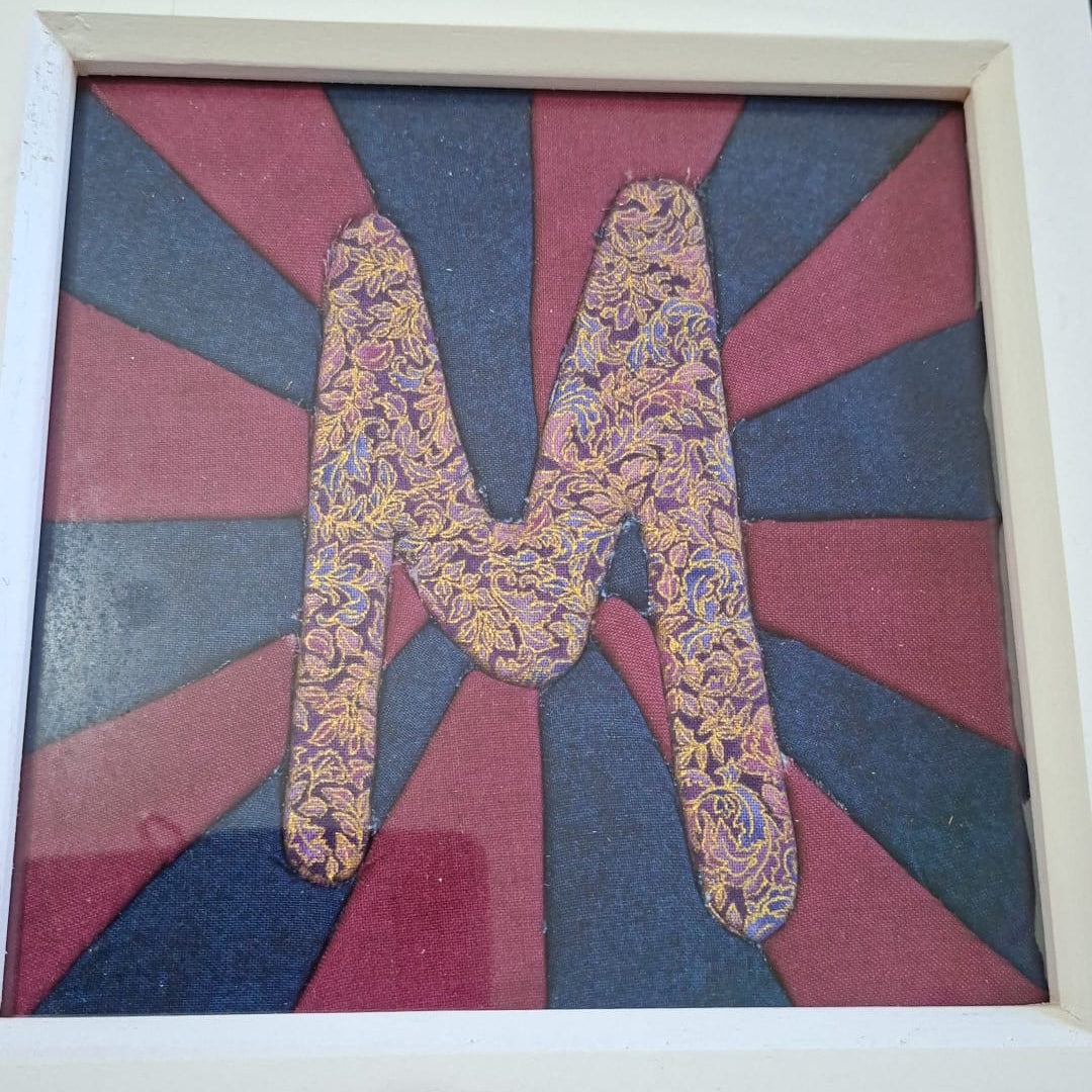 A framed decorative art piece featuring a large textured letter 'M' in a multicolor floral pattern, set against a background of alternating red and blue sunburst rays. The piece is housed in a white wooden frame and photographed on a green cutting mat with craft supplies in the background.